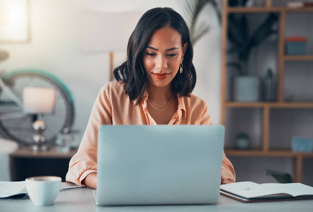 A woman with shoulder-length hair is smiling while working on a laptop in her home office, possibly drafting a government report. She is wearing a peach-colored blouse. A bicycle and shelves with decor are in the background. A notebook and a cup are on the desk.